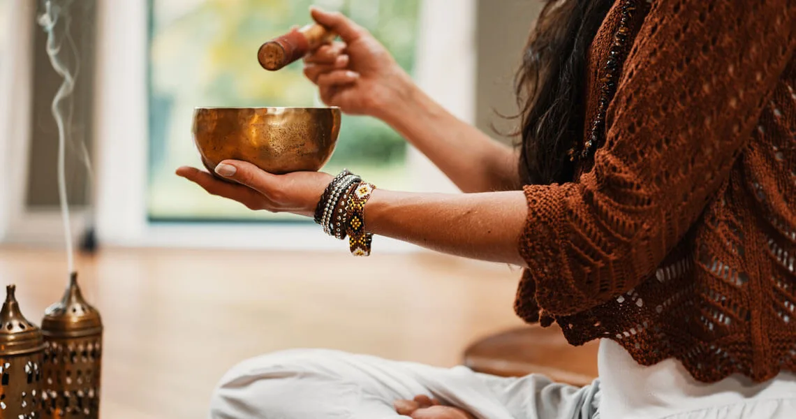 Femme de profil portant un pull marron clair, tenant un bol doré dans les mains, bracelets multicolores au poignet, intérieur lumineux avec fenêtre et objets de décoration dorés en arrière-plan.