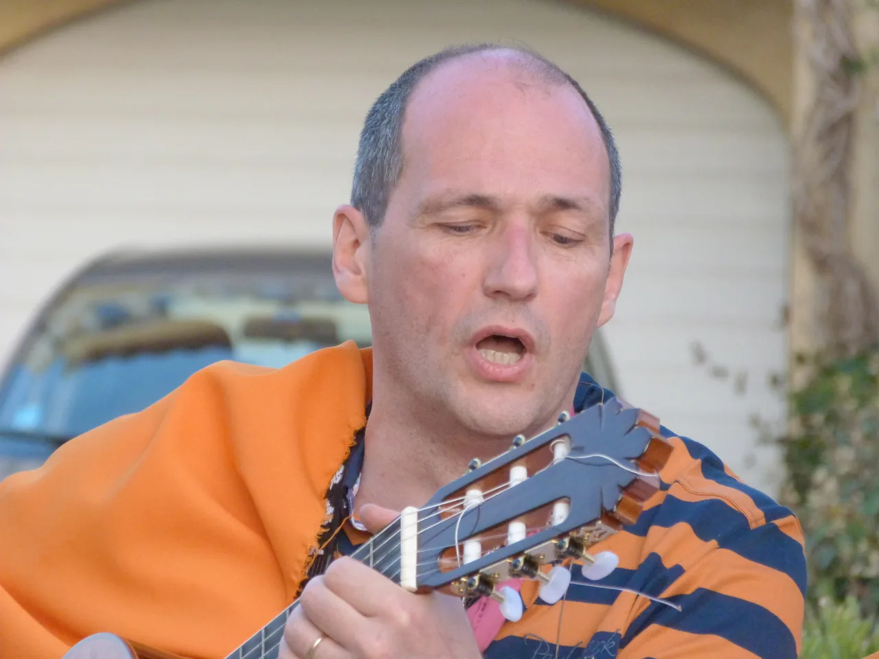 Homme chauve en chemise orange et pull bleu marine tenant une guitare classique, bouche ouverte comme chantant, devant mur blanc avec voiture bleue en arrière-plan.
